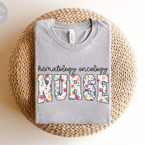 Puede incluir: Una camiseta gris con el texto "hematology oncology NURSE" en letras blancas con un patr&oacute;n floral. La camiseta est&aacute; sobre una superficie de paja tejida.
