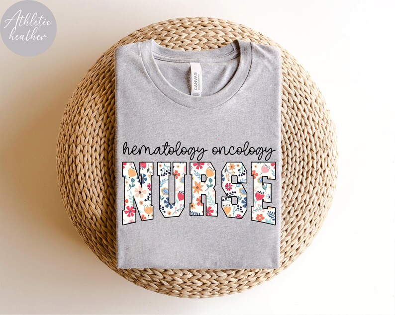 Puede incluir: Una camiseta gris con el texto "hematology oncology NURSE" en letras blancas con un patr&oacute;n floral. La camiseta est&aacute; sobre una superficie de paja tejida.