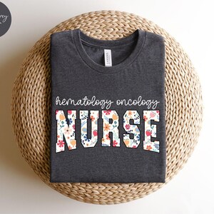 Puede incluir: Camiseta de color gris oscuro jaspeado con un dise&ntilde;o floral que dice "hematology oncology nurse".