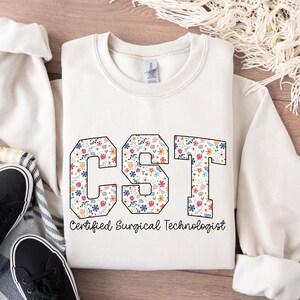 May include: A white sweatshirt with the text "CST Certified Surgical Technologist" printed on the front. The letters are outlined in black and filled with a floral pattern in shades of pink, orange, blue, and green.