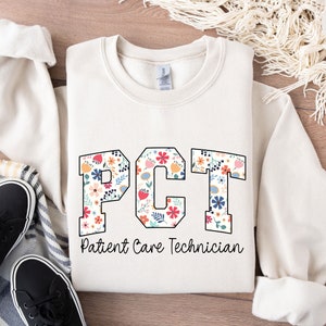 Puede incluir: Una sudadera blanca con un diseño de estampado floral que deletrea "PCT" para Patient Care Technician.