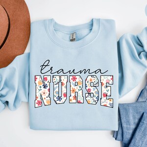 K&ouml;nnte beinhalten: Hellblauer Pullover mit dem Text "trauma nurse" in wei&szlig;en umrandeten Buchstaben. Die Buchstaben sind mit einem floralen Muster in den Farben Rosa, Rot, Gelb und Blau gef&uuml;llt.