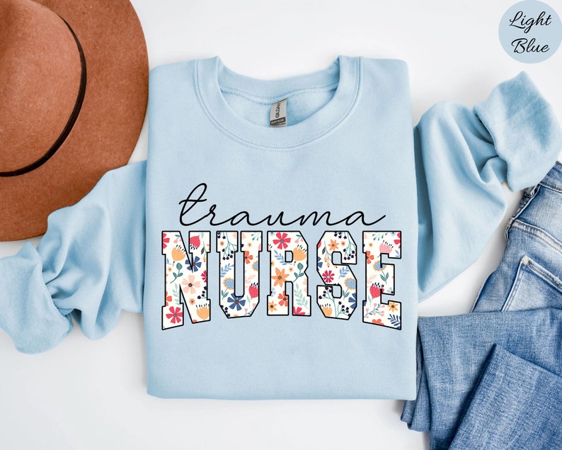 K&ouml;nnte beinhalten: Hellblauer Pullover mit dem Text "trauma nurse" in wei&szlig;en umrandeten Buchstaben. Die Buchstaben sind mit einem floralen Muster in den Farben Rosa, Rot, Gelb und Blau gef&uuml;llt.