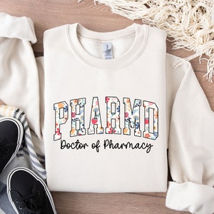 May include: A cream-colored sweatshirt with a floral print design that says "PHARM.D. Doctor of Pharmacy".