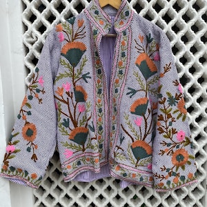 Puede incluir: Chaqueta lavanda con bordados florales en tonos naranja, verde y rosa. La chaqueta presenta un patrón de espiga y un forro a rayas. El bordado es detallado, creando un aspecto texturizado. La chaqueta está colgada en una percha de madera.