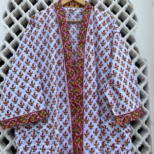 Puede incluir: Bata de algodón blanca con un estampado floral rojo y amarillo repetido. La bata tiene un borde floral rojo y amarillo contrastante a lo largo de los bordes y las mangas. La bata está colgada de una percha de madera.