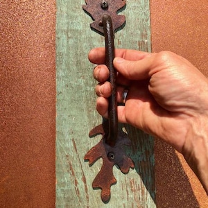 May include: A dark brown, rusted metal door handle featuring decorative oak leaf-shaped mounting plates at the top and bottom. The handle is secured with visible screws to a weathered, light green painted wooden board with a distressed finish.