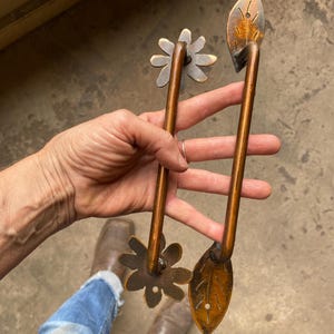 May include: Two metal door handles with a warm bronze finish. One handle features a flower-shaped mounting plate at each end, while the other handle features a leaf-shaped mounting plate at each end, each with a small hole for mounting hardware.