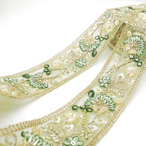 Può includere: Primo piano di un bordo decorativo con un design floreale. Il bordo ha una base trasparente verde chiaro con fiori ricamati in tonalità di verde e crema, accentuati da filo d'oro e piccole perline.