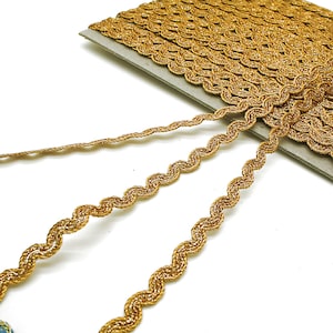 May include: Close-up of gold metallic trim with a wavy, scalloped edge. The trim is tightly woven and appears to be on a roll. The trim is a bright, shiny gold color. The trim is likely used for sewing, crafting, or embellishing clothing and accessories.