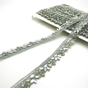 May include: A roll of silver-gray decorative trim with a repeating pattern of clear and gold-toned embellishments. The trim is made of a woven material and features a delicate, ornate design, perfect for embellishing clothing or crafts.
