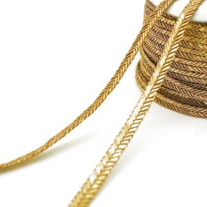 May include: Close-up of a spool of gold-colored braided trim. The trim has a herringbone pattern and is tightly wound. A length of the trim extends from the spool, showcasing its detailed weave and metallic sheen. The background is plain white.