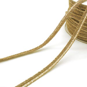 Może przedstawiać: Złota ozdobna lamówka na szpuli. Lamówka ma pleciony wzór i jest pokazana na białym tle. Lamówka jest prawdopodobnie używana do szycia, rękodzieła lub ozdabiania odzieży i akcesoriów.