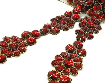 May include: A roll of decorative trim featuring ruby red faceted glass stones. The stones are arranged in a floral pattern and are connected by a gold-colored wire. The trim is on a white background.
