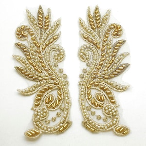 May include: Two ornate, symmetrical gold beaded appliques on a white mesh backing. The designs feature a combination of seed beads and larger gold beads, creating a textured, floral-inspired pattern. The appliques are likely for embellishing garments or accessories.