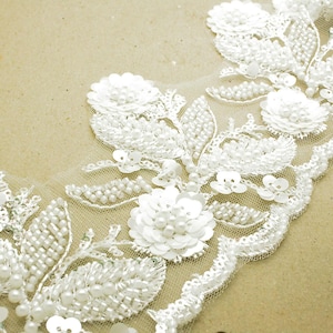 May include: Close-up of white lace trim with floral and leaf designs. The trim features beaded accents and sequin details, creating a delicate and textured appearance. The lace is set against a neutral background.
