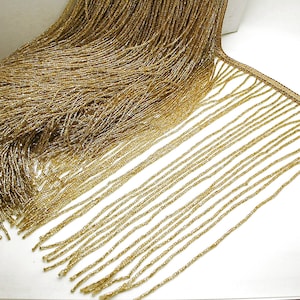 May include: Gold beaded fringe trim. The trim features a dense top section of small, gold beads, transitioning into long, dangling strands. The beads have a metallic sheen, creating a shimmering effect. This trim is suitable for embellishing clothing, accessories, or home decor projects.