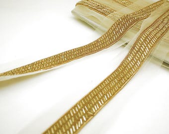 Puede incluir: Ribete de cuentas doradas sobre una tela transparente blanca. El ribete presenta un patrón repetitivo de pequeñas cuentas doradas rectangulares. El ribete se muestra en primer plano, destacando los intrincados detalles y la textura del diseño. El ribete se utiliza probablemente para embellecer ropa o manualidades.