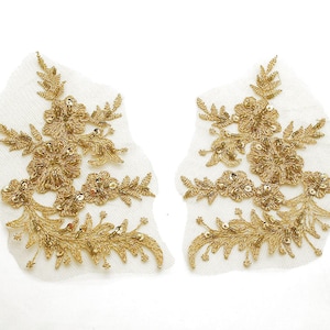 以下が含まれることがあります： 白いメッシュの背景に、2つの金色の刺繍の花のアップリケ。アップリケは、詳細な金色の糸細工、スパンコール、繊細な花の模様が特徴です。衣服やクラフトプロジェクトの装飾に最適です。