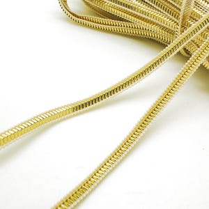 May include: A spool of metallic gold trim featuring a central raised ridge with a repeating rectangular segment pattern, bordered by thin, textured cream-colored piping along both edges, presented in a coiled arrangement on a white surface.