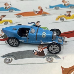 May include: A small, light blue toy race car with the number 8 on the side. The car has black wheels and silver accents. The background features a repeating pattern of cartoon race cars in various colors.