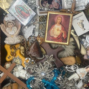Può includere: Una collezione di oggetti religiosi, tra cui immagini incorniciate, croci e collane di perline. Le immagini raffigurano figure e scene religiose. Sono visibili vari colori e trame, tra cui oro, argento e legno.