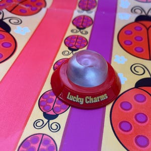 Könnte beinhalten: Ein rotes und silbernes Lucky Charms Spielzeug mit dem Aufdruck "Lucky Charms" an der Seite. Das Spielzeug befindet sich auf einem farbenfrohen Hintergrund mit Marienkäfer- und Blumendesigns in Rot, Rosa und Lila.
