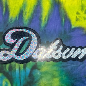 May include: A sticker with the word "Datsun" in a stylized font. The word is outlined in black with a holographic, iridescent pattern. The background is a tie-dye pattern in shades of blue, green, and yellow.