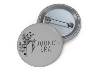 May include: A gray button with a black and white illustration of a book with flowers coming out of it. The text "BOOKISH ERA" is printed on the button.