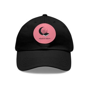 Pode incluir: Boné de beisebol preto com um patch circular rosa com uma lua crescente, um livro e o texto "Bookish Babe".