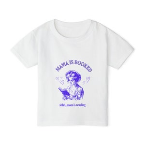 Puede incluir: Camiseta blanca con un gráfico morado de una mujer leyendo un libro. El texto "Mama is booked" está por encima de la imagen y "shhh...mom is reading" está por debajo de la imagen.