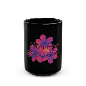 Könnte beinhalten: Schwarze Keramiktasse mit einem Blumendesign und dem Text "Persephone's Garden Club" in Rosa.