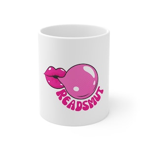 以下が含まれることがあります： ピンクのガムと唇のデザインが施された白いセラミックマグカップ。ガムの下にピンク色の文字で「READSMUT」と書かれています。