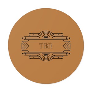 Könnte beinhalten: Ein runder, brauner Untersetzer mit einem schwarzen Art-Deco-Design. Das Design zeigt ein rechteckiges Banner mit den Buchstaben "TBR". Der Untersetzer besteht aus einem strapazierfähigen Material.