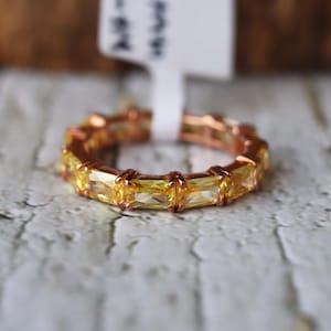 Könnte beinhalten: Ein goldfarbener Ring mit einer Reihe gelber, rechteckiger Edelsteine. Der Ring liegt auf einer hellen Oberfläche, mit einem Etikett im Hintergrund. Die Edelsteine sind gleichmäßig um das Band angeordnet.