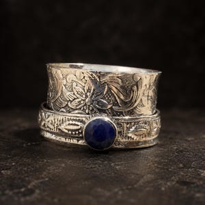 May include: A wide silver spinner ring featuring an intricate floral and scrollwork pattern on the upper band. The lower band displays a textured leaf motif and is set with a single, round, faceted dark blue gemstone centered on the front.