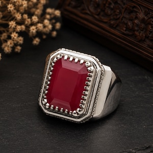 May include: A silver ring featuring a large, rectangular, deep red gemstone set in a pronged bezel. The metal band has a polished finish, and the stone is surrounded by a decorative, notched frame that adds texture to the overall design of the piece.