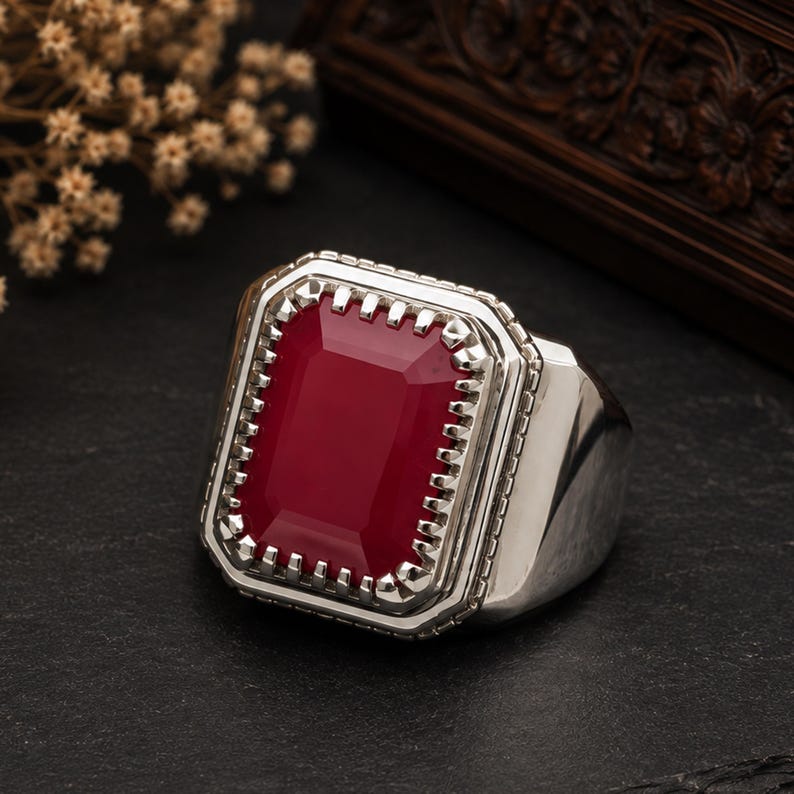 May include: A silver ring featuring a large, rectangular, deep red gemstone set in a pronged bezel. The metal band has a polished finish, and the stone is surrounded by a decorative, notched frame that adds texture to the overall design of the piece.