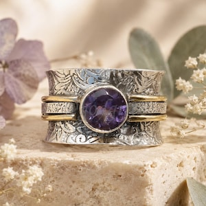 May include: A wide silver band ring featuring an intricate floral embossed pattern and a central round faceted purple gemstone. The ring includes two thin, smooth gold-toned metal bands that wrap horizontally around the center of the silver band.