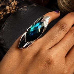 Può includere: Un anello importante con una grande pietra di labradorite taglio marquise dalla superficie sfaccettata che mostra riflessi iridescenti blu e verdi. La pietra è incastonata in una fascia in metallo color argento lucido che si restringe alle estremità.