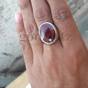 Puede incluir: Un anillo de plata con una gema roja oscura grande, de corte ovalado. El anillo está engastado en una banda de plata pulida y sencilla. La gema tiene múltiples facetas, que reflejan la luz.