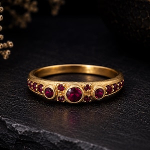 Puede incluir: Un anillo de oro con acabado mate y texturizado que presenta un rubí rojo central de talla redonda, flanqueado por rubíes más pequeños de talla redonda engastados a lo largo de la banda. El diseño incorpora gemas rojas de varios tamaños.