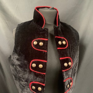 May include: A black velvet vest with a red trim and silver buttons. The vest has a mandarin collar and a cropped length.