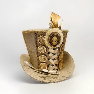 May include: A tan sequined top hat with gold detailing. The hat is adorned with a cream cameo, gold leaf accents, and decorative gold buttons. A cream ribbon laces up the front of the hat.