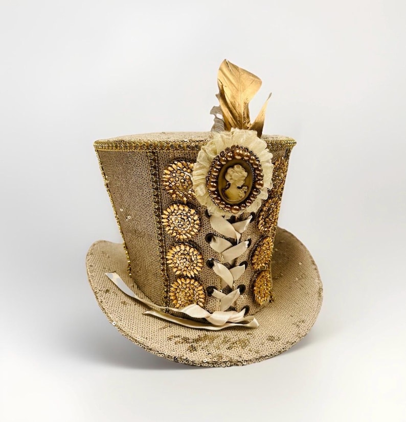 May include: A tan sequined top hat with gold detailing. The hat is adorned with a cream cameo, gold leaf accents, and decorative gold buttons. A cream ribbon laces up the front of the hat.