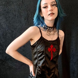 May include: A woman with blue hair and dark eyeliner wears a black satin tank top with a red cross design. She is wearing a silver choker necklace with multiple chains.