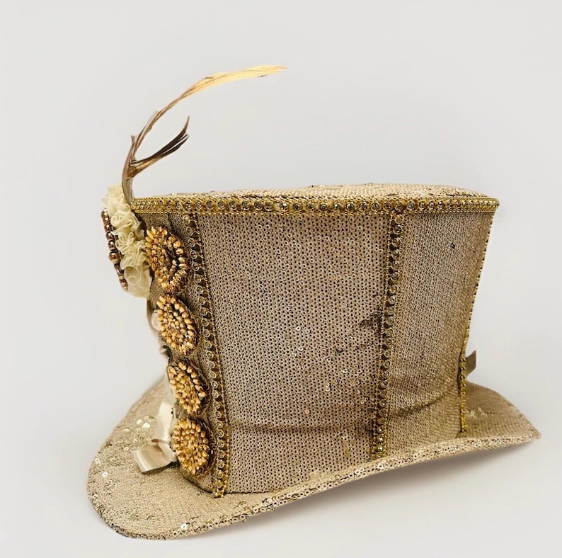 May include: A gold sequined top hat with decorative embellishments. The hat features a small antler and feather, along with beaded floral accents and a satin ribbon. The hat is a light tan colour.