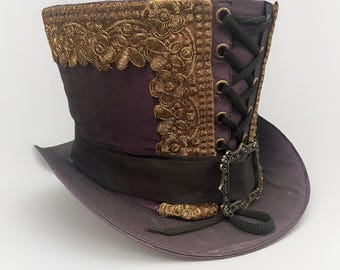 Può includere: Un cappello a cilindro viola con tesa e fascia nere. Il cappello presenta ricami floreali dorati e un design allacciato nero sul retro. Una fibbia decorativa e dettagli dorati completano il look.