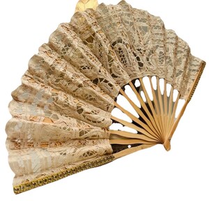 May include: A beige lace hand fan with a wooden frame. The fan is open, showing a delicate, patterned lace design. The edges are trimmed with gold-coloured embellishments. The fan is a decorative accessory.