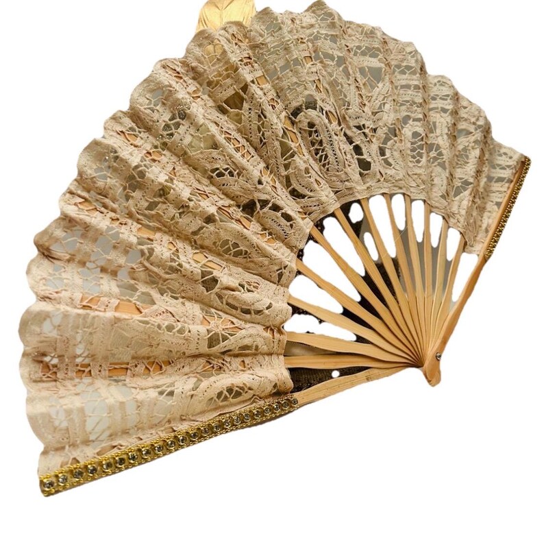 May include: A beige lace hand fan with a wooden frame. The fan is open, showing a delicate, patterned lace design. The edges are trimmed with gold-coloured embellishments. The fan is a decorative accessory.
